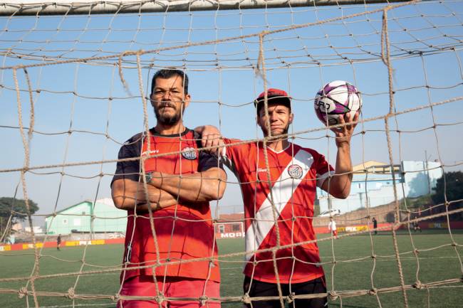 Imagem mostra pai. Dirceu, e filho, Alberto, em campo de futebol. Os dois estão dentro de gol e olham para câmera por entre as redes do gol. O filho segura no braço esquerdo uma bola de futebol.