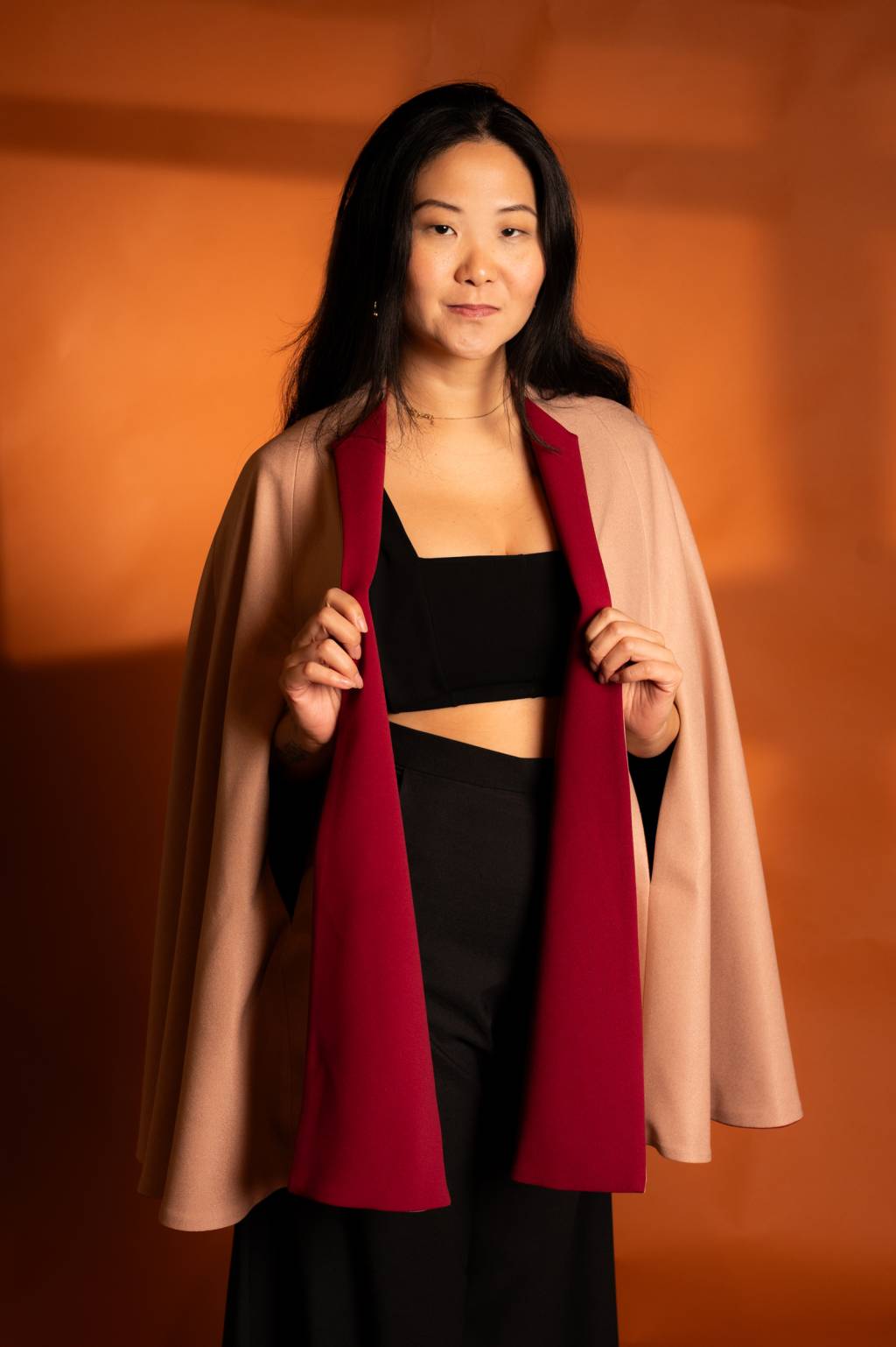 Julia Pak (Thiago Farias-Divulgação) (2) Mulher asiática de cabelos pretos lisos longos sorri segurando seu casaco nas cores vinho e bege. O fundo é laranja.