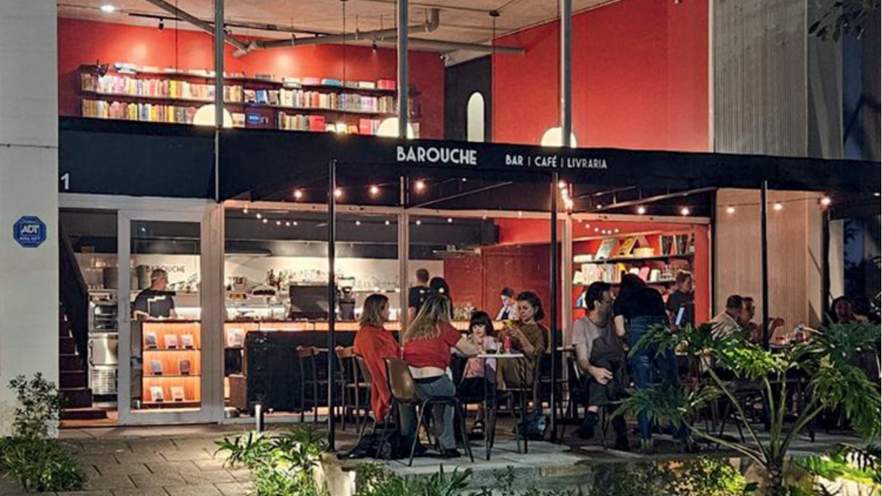 A imagem mostra a fachada do Barouche. Tem mesas do lado de fora, na calçada, com pessoas. No interior do bar, as paredes são vermelhas.