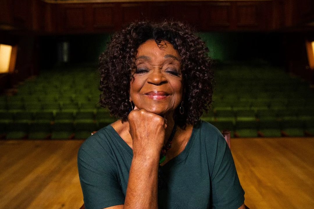 uma mulher negra com os cabelos soltos na altura do ombro. ela esta sentada, sorrindo e apoiando o rosto na mão direita.