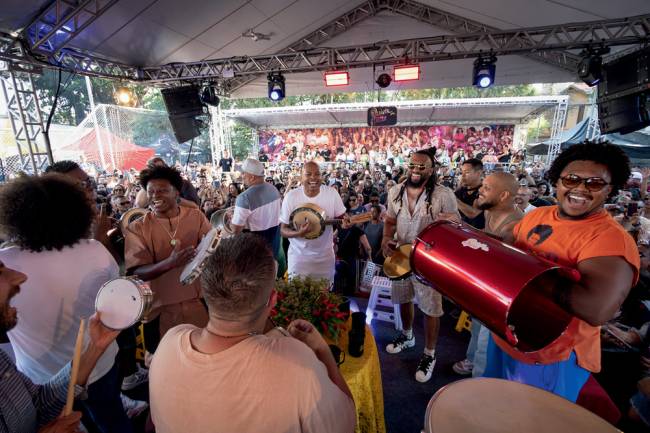 Quintal dos Prettos Em primeiro plano, imagem mostra roda de samba, com vários homens tocando. Ao fundo, multidão em ambiente parcialmente aberto