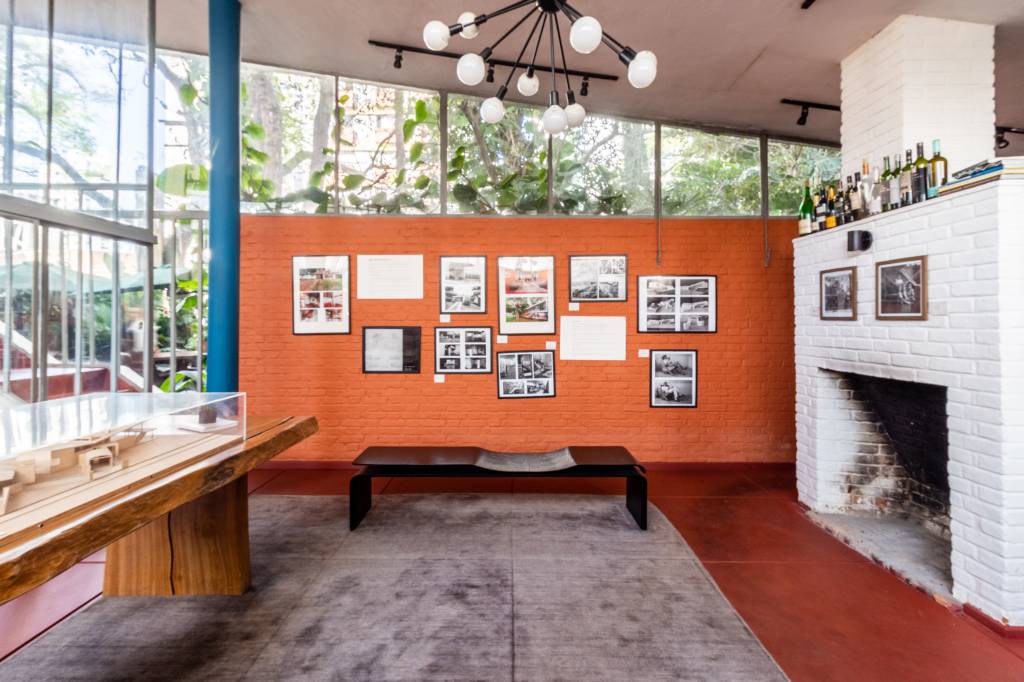 Segunda Residência de Vilanova Artigas – Campo Belo – 1949 – @o Parede laranja, teto alto, grande lustre, móveis de madeira, quadros e tapete cinza aparecem em destaque em sala de estar.