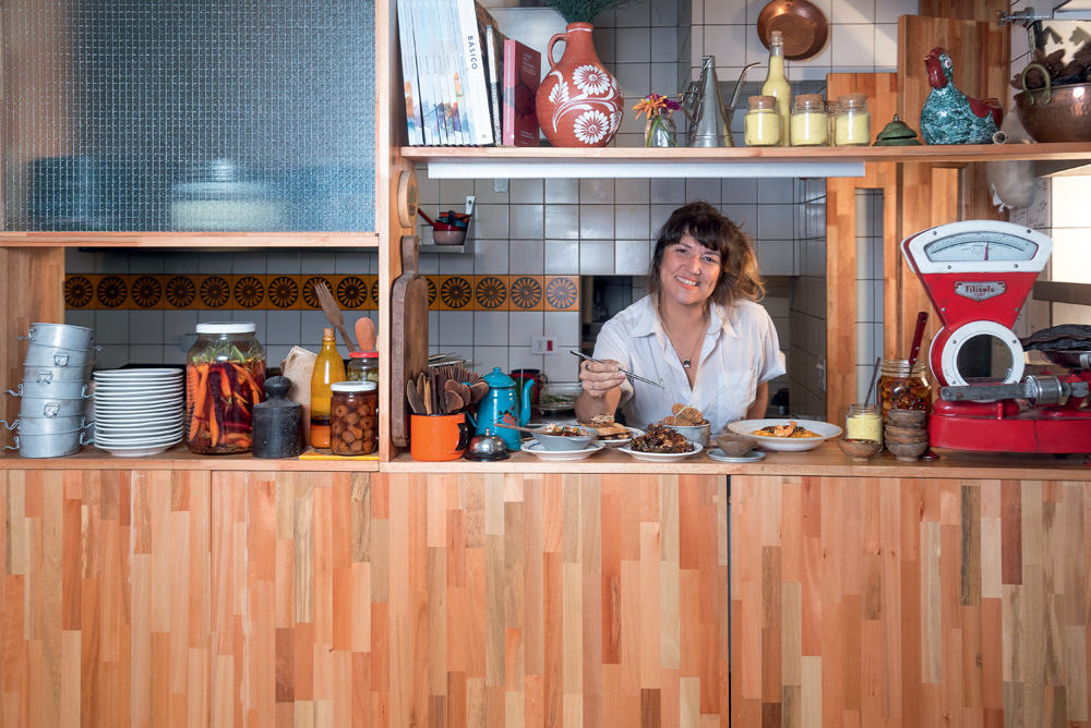 COMER & BEBER 2023 – Aconchegante Bar A imagem mostra uma mulher atrás de um balcão de madeira, ela sorri para a foto. No balcão a sua frente tem seis pratos com comidas. À esquerda no balcão tem uma pilha de pratos, panelas e potes de tempero.