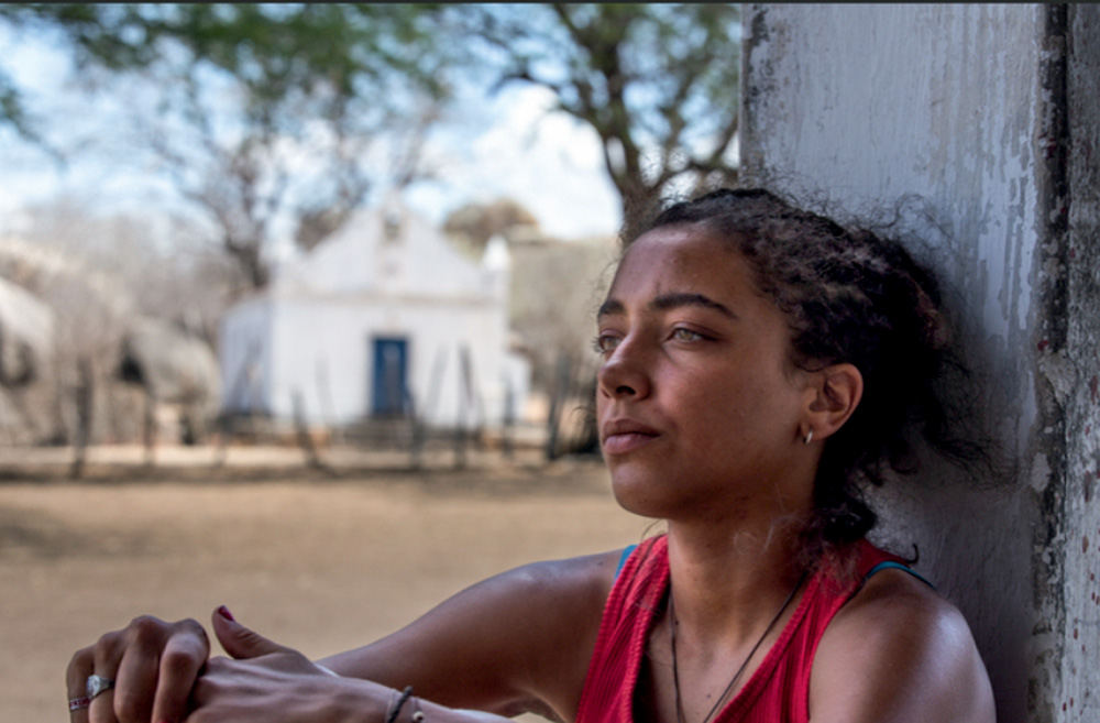Cangaço Novo (Amazon Studios-Divulgação) (1).jpg Mulher de pela morena aparece encostada em parede branca, sentada com olhar distante. Ao fundo, uma pequena igreja.