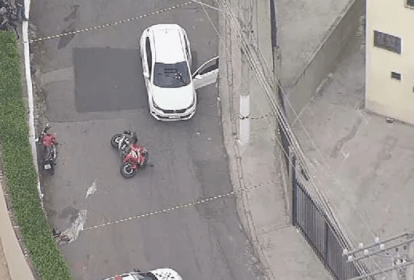 a imagem mostra um carro branco com marcas de tiro no para-brisa e, ao lado, uma moto caída no chão