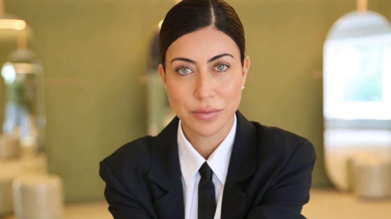 Mulher branca de cabelos lisos presos posa de pernas cruzadas com terno preto e camisa branca.