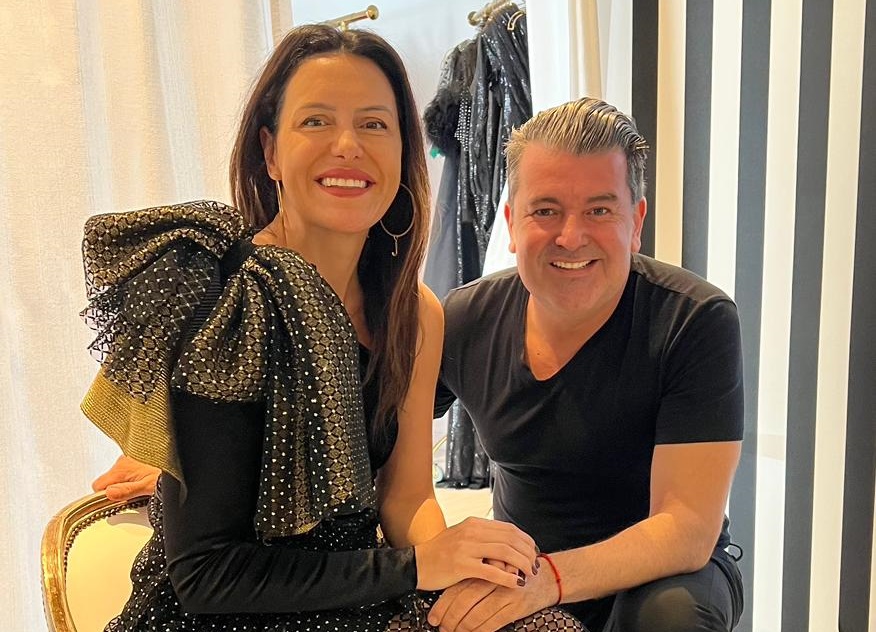 Mulher sentada em cadeira dourada sorri ao lado de homem grisalho de joelhos. Ela veste vestido preto com saia transparente e detalhes em um dos ombros. Ele veste camiseta e calça pretas.