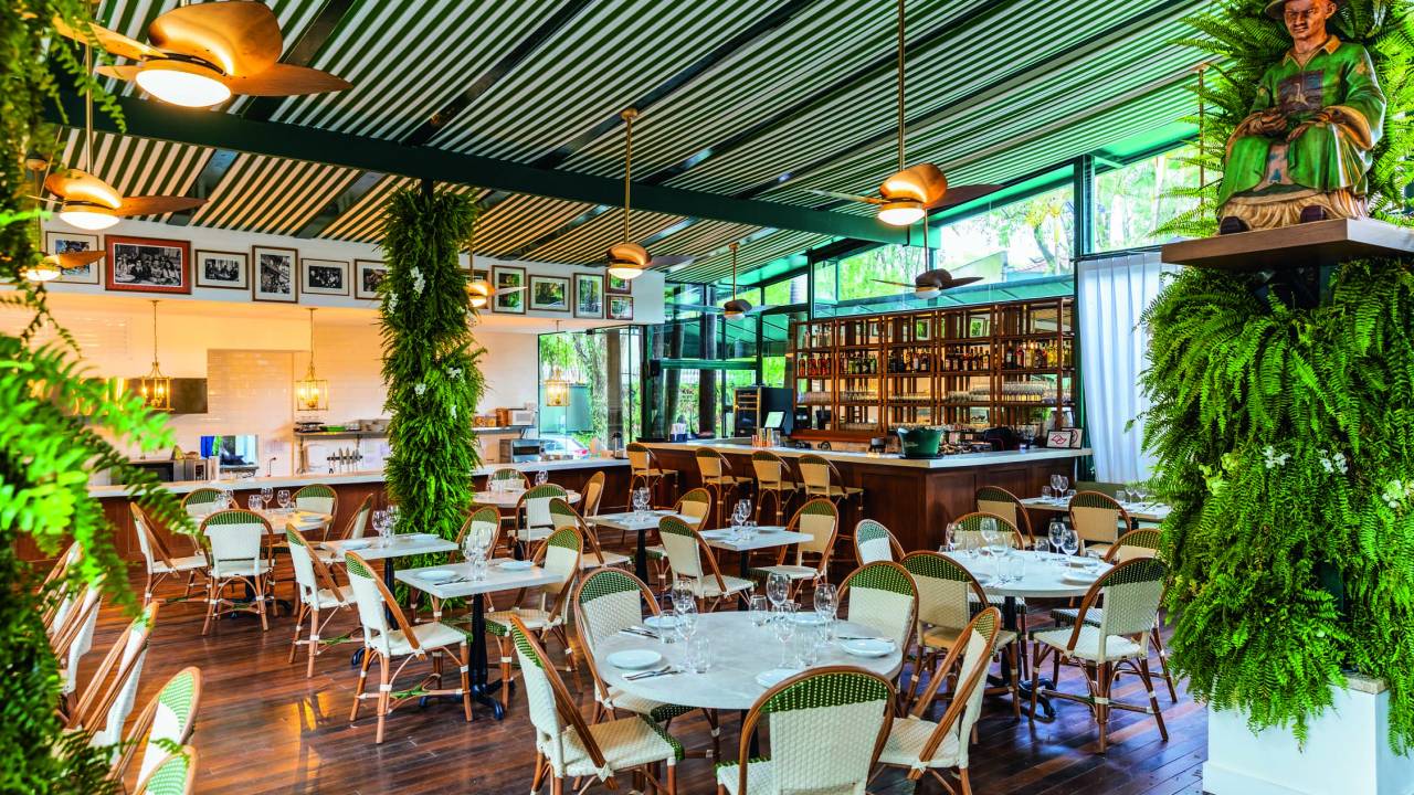 Salão de restaurante com chão de madeira, mesas de madeira com branco e bar ao fundo direito.