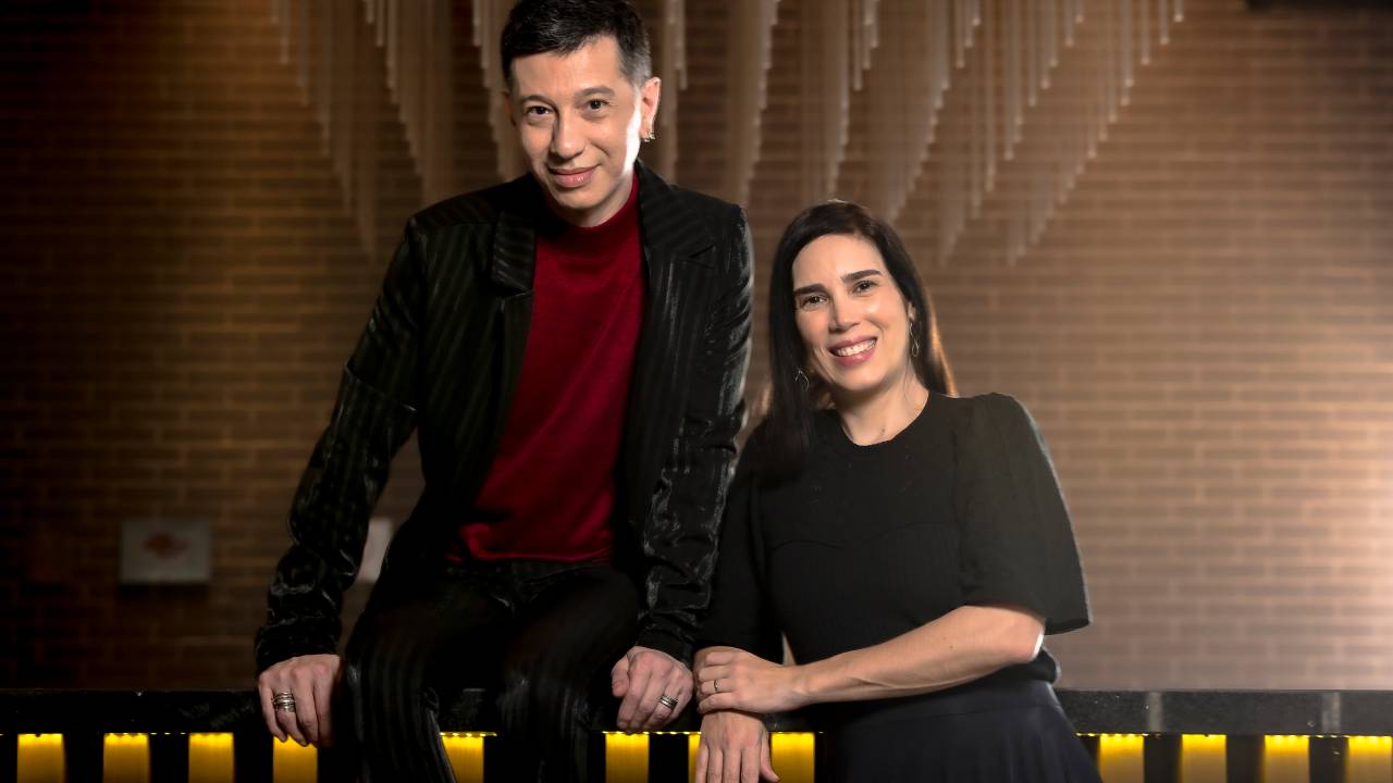 Homem branco posa de pé, apoiado em batente dourado atrás. Veste blazer preto com suéter cor de vinho. Ao lado, mulher branca de cabelos longos posa sentada com vestido preto.
