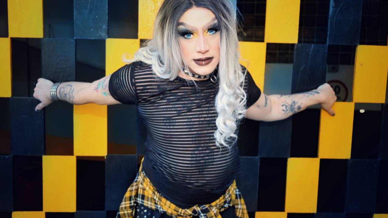 Drag queen posa com os braços na parede quadriculada, usa peruca de cabelo loiro longo e veste camiseta preta transparente com jaqueta quadriculada na cintura.