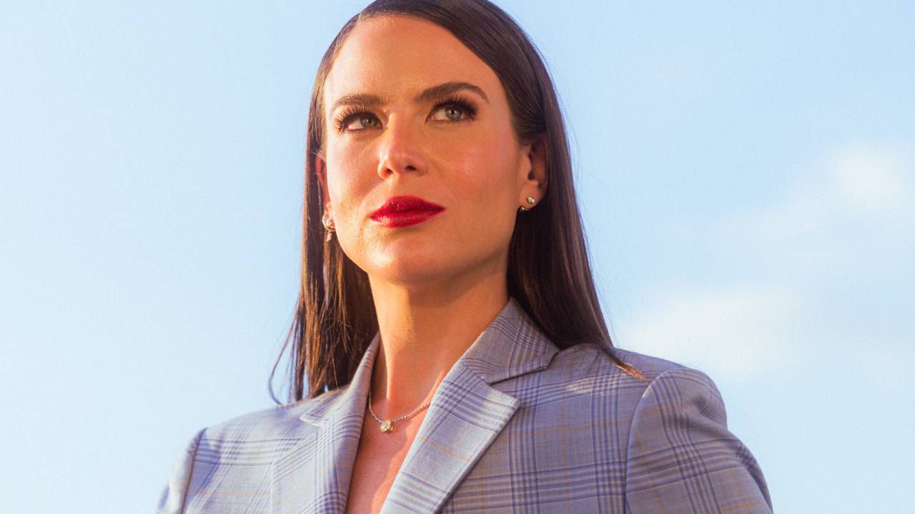 Mulher branca com cabelo bem liso e longo posa olhando para o horizonte, com as mãos na cintura. Veste blazer azul claro e o céu azul diurno aparece ao fundo.