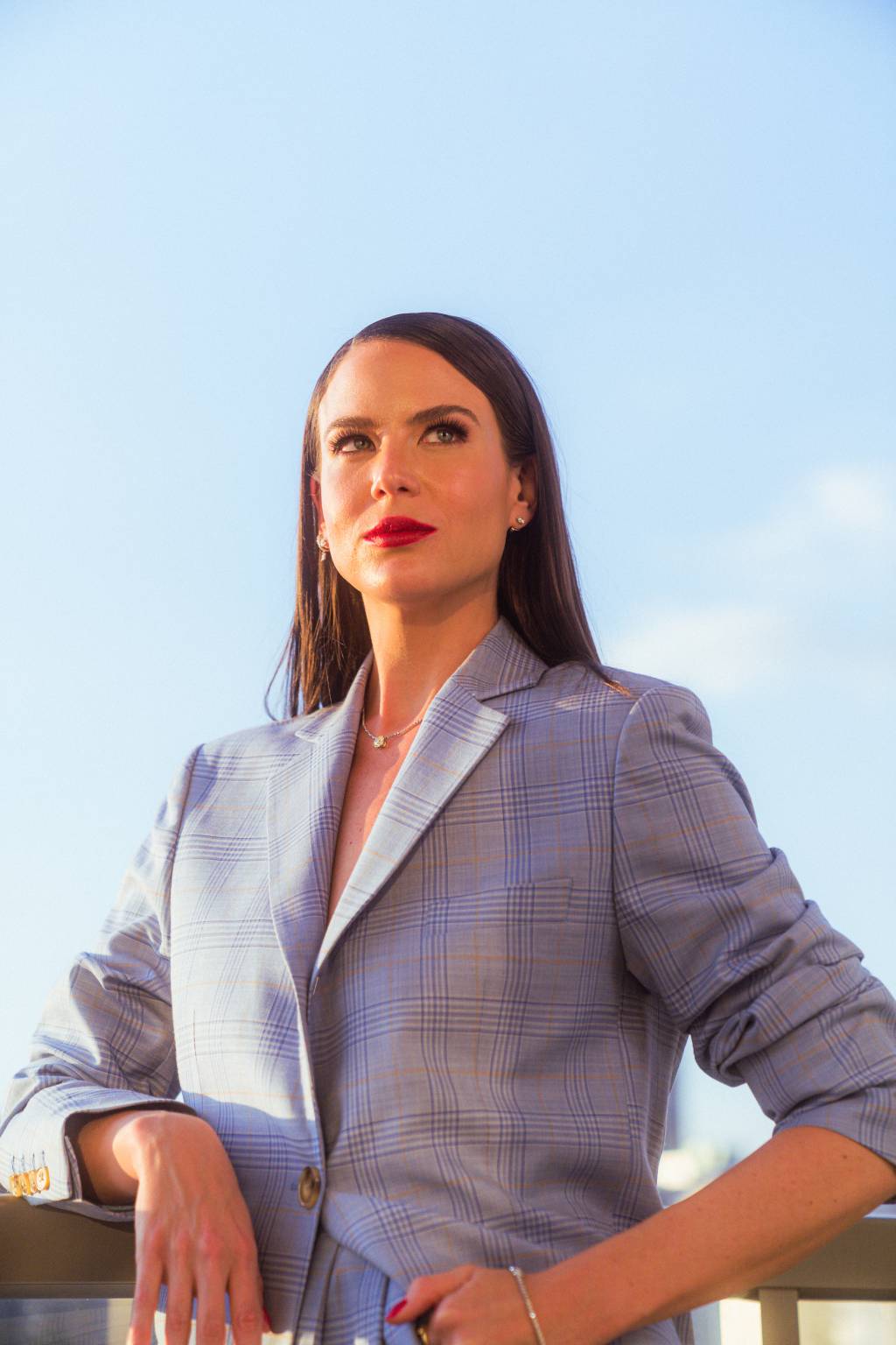 Nathalie Felsberg – willian soares Mulher branca com cabelo bem liso e longo posa olhando para o horizonte, com as mãos na cintura. Veste blazer azul claro e o céu azul diurno aparece ao fundo.