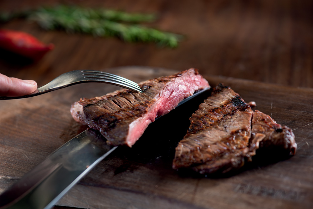 COMER & BEBER 2023 – Rubaiyat Um pedaço de carne cortada ao meio com uma faca por baixo e um garfo fincado nela.
