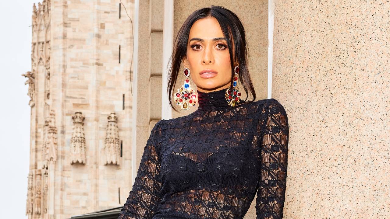 Mulher branca e magra posa encostada em pilar branco. Veste roupa toda preta com transparência que exibe parte de baixo, também preta.