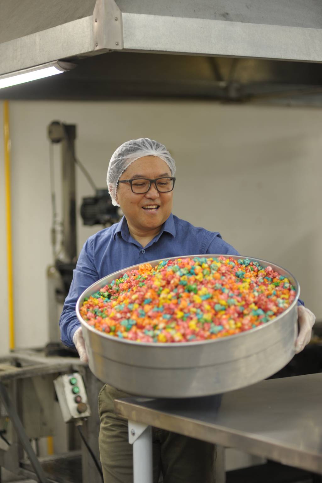 Toshio Hito (Angélica Covo-Divulgação) Homem asiático de camisa social azul e proteção no cabelo segura grande panela repleta de pipocas coloridas.