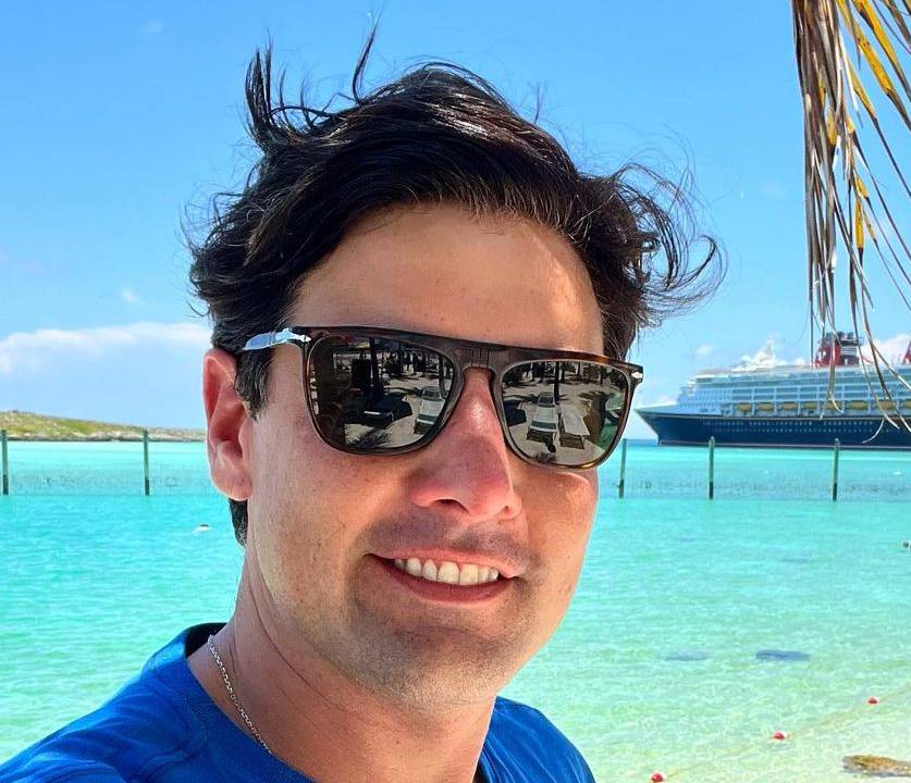 bruno de luca usa óculos de sol em uma praia e, ao fundo, há um navio