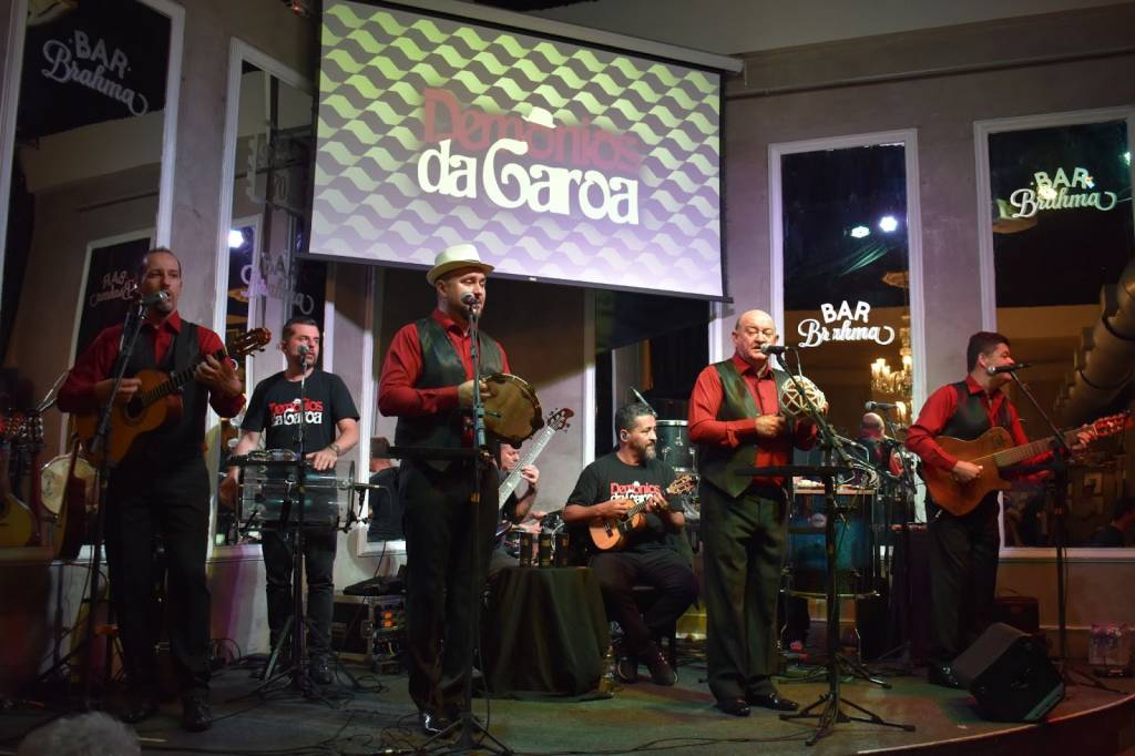 Bar Brahma – Demônios da Garoa Pequeno palco do bar com sete músicos. Os quatro da frente estão em pé e vestem camisa vermelha com colete preto. Tocam cavaquinho, pandeiro, chocalho e guitarra. Ao fundo, três homens tocam as percussões.