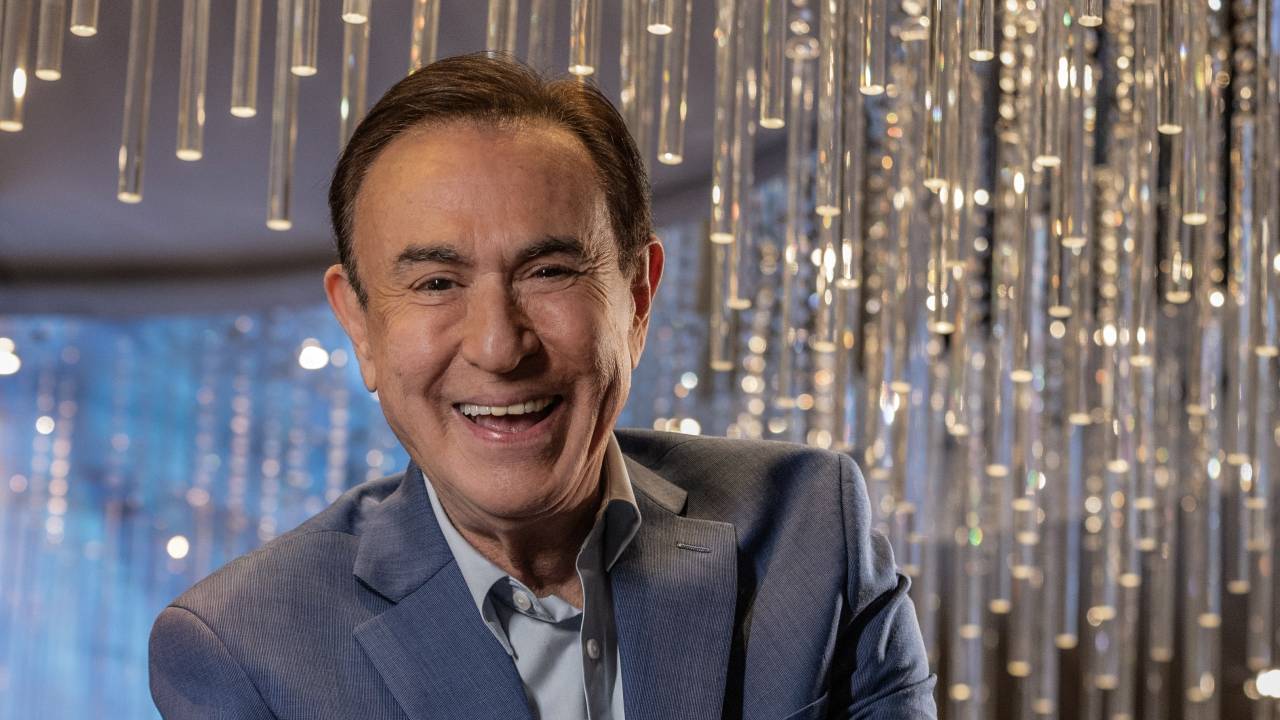 Homem branco de terno e gravata azuis e peças longas iluminadas de cristal ao fundo.