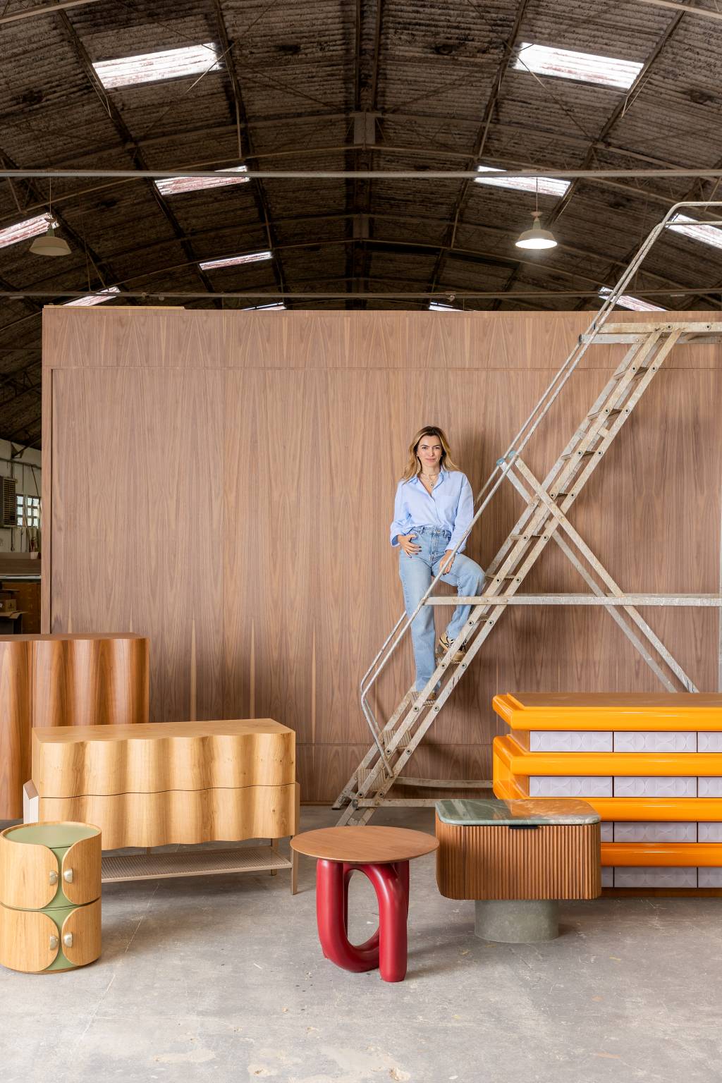Ana Weege (Rafael Renzo-Divulgação) Mulher loira posa de pé em grande escada em um galpão com vários móveis coloridos espalhados ao redor. Ela veste camisa e calça jeans.