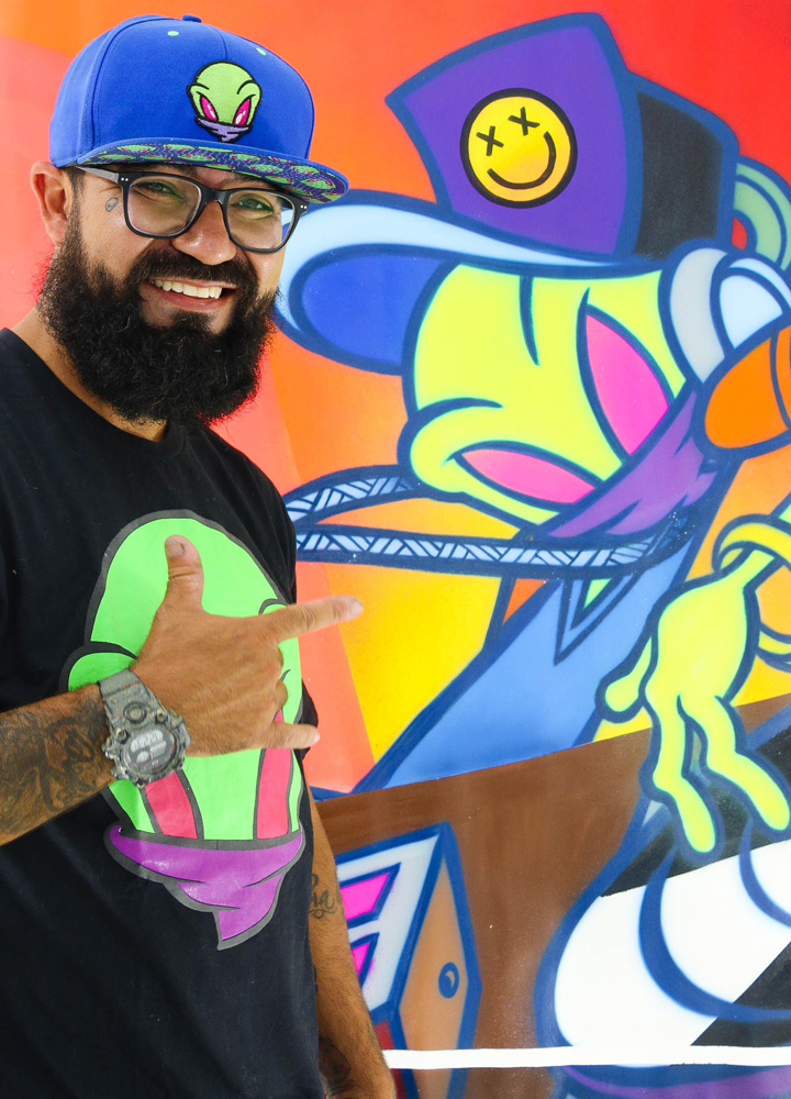 André Gomes Romão (Caroline Paixão-Divulgação).jpeg Homem branco de barba grande, óculos e boné posa com uma mão erguida em frente a parede de grafite colorido com um extraterrestre de boné.