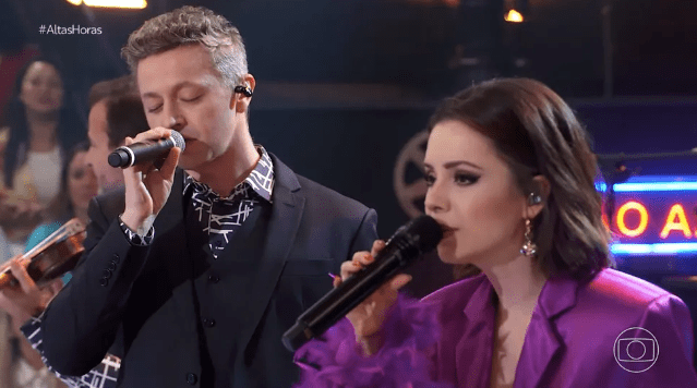 um homem branco de roupa social e cabelos curtos cantando de olhos fechados ao lado de uma mulher branca, de cabelos castanhos, lisos, na altura do ombro, e vestido roxo brilhante também cantando