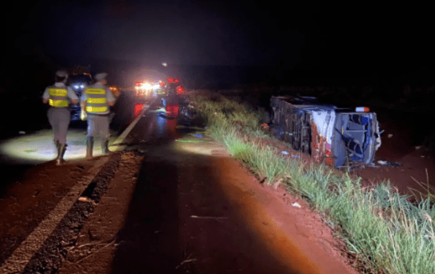 policiais militares andando pela estrada à noite e um ônibus capotado