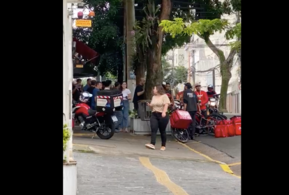 Frame de vídeo mostra pessoas passando na calçada de rua. Ao fundo, parcialmente encoberto por árvores, entregadores discutem com homem branco