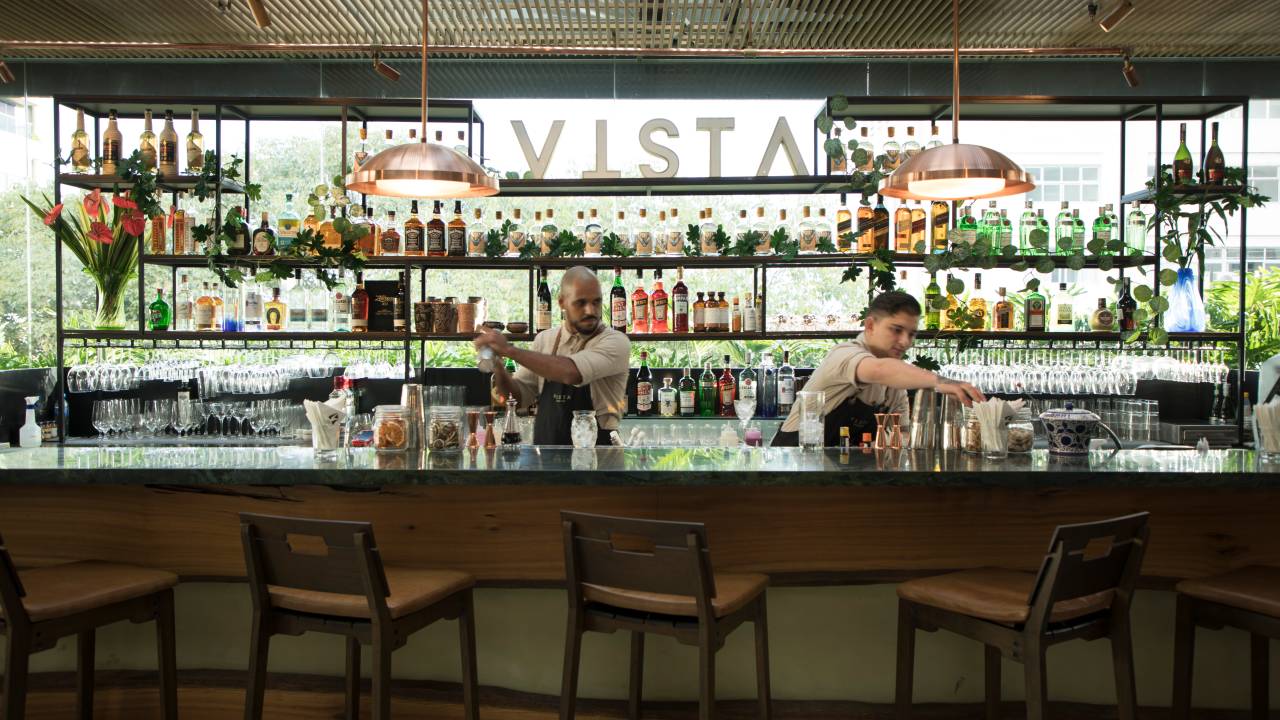 Balcão do bar do restaurante vista com cadeiras altas de madeira na frente, dois bartenders trabalhando e uma parede cheia de garrafas de bebida atrás.