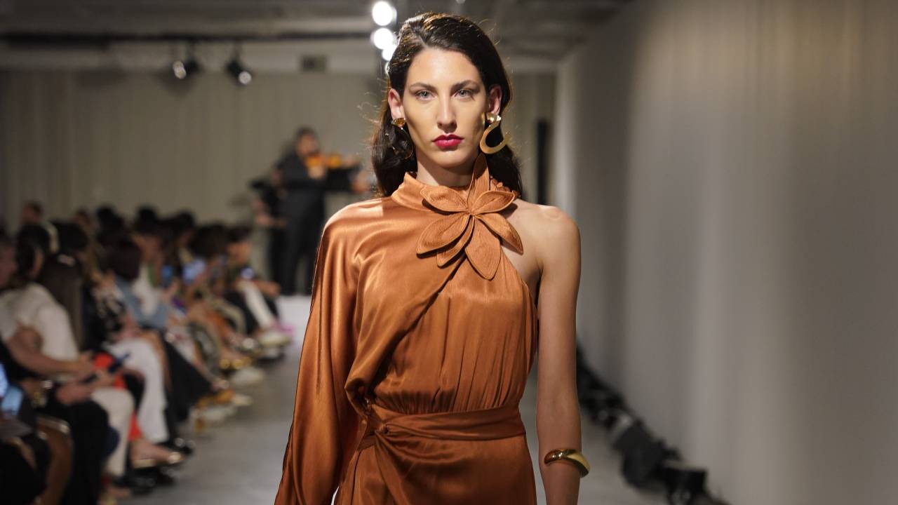 Foto exibe modelo branca andando em passarela de chão cinza com fileira de pessoas sentadas à esquerda. Ela veste um vestido de cor marrom claro que cobre todo seu torso e exibe parte de uma das pernas.