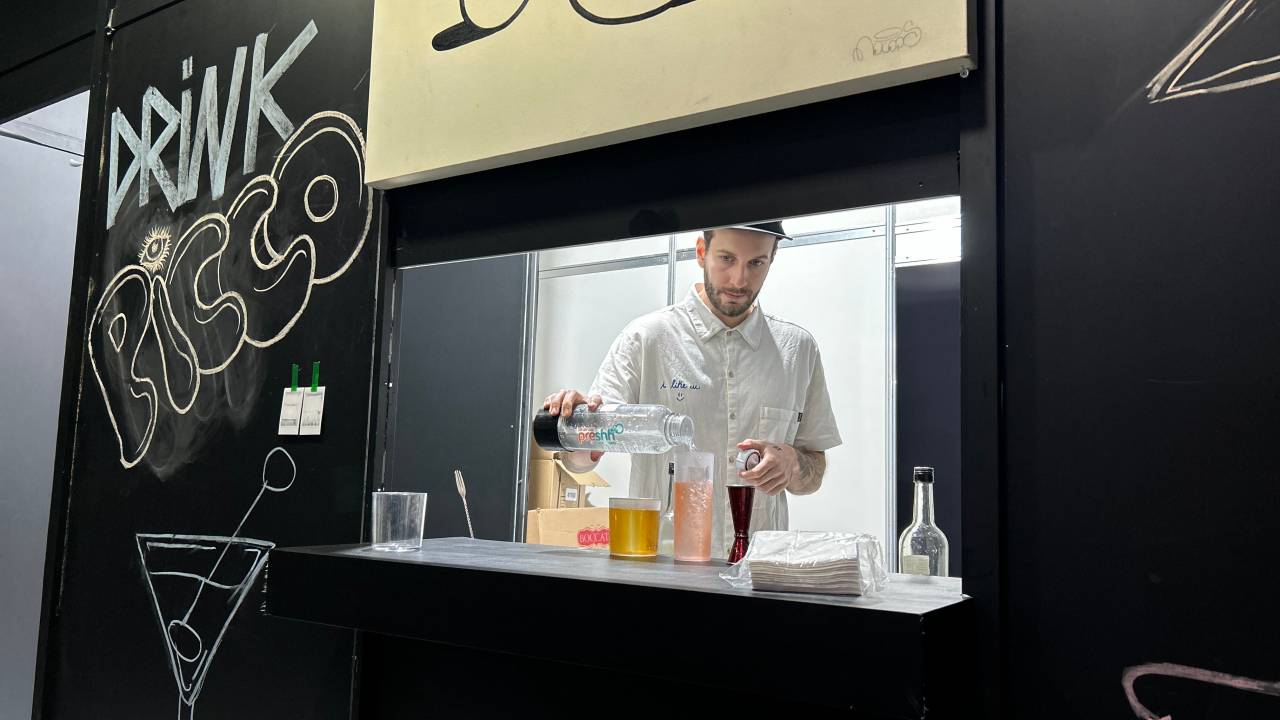 Homem atrás de janela em estande de paredes pretas preparando drinques coloridos.