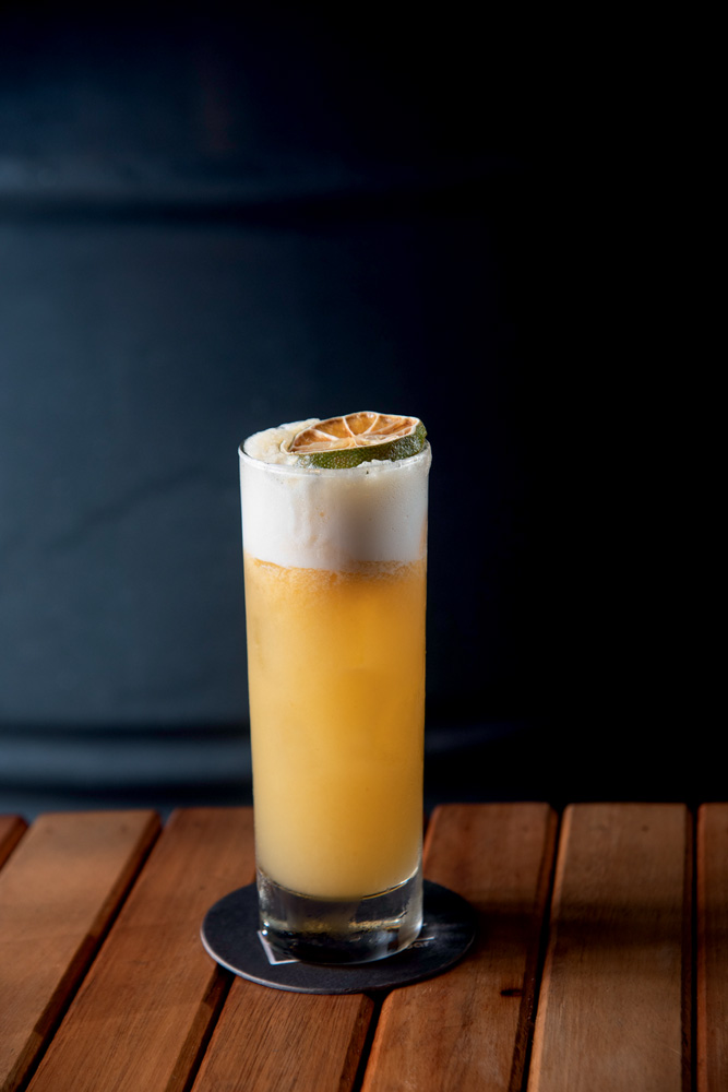 Lupe Bar Sobre uma mesa de madeira, um copo de vidro com drinque na cor alaranjada. No topo do copo tem bastante espuma e uma rodela de laranja.