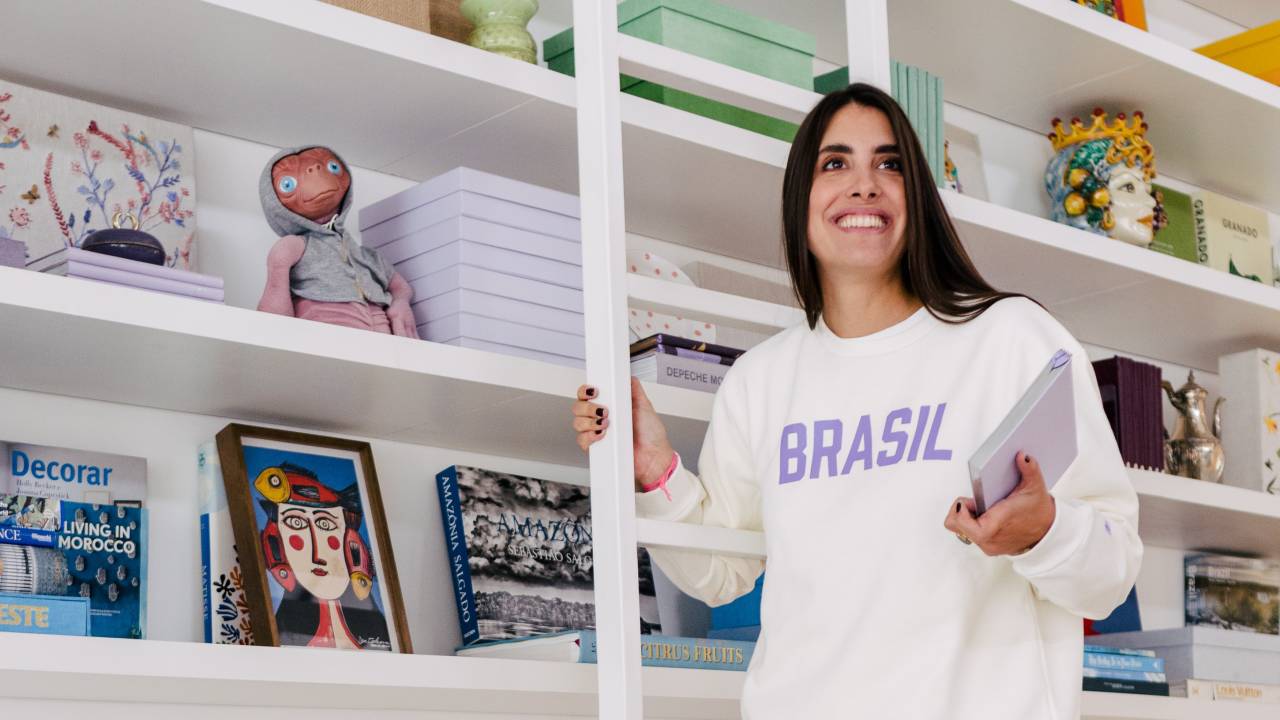 Mulher de cabelo liso escorrido preto sorri de pé ao lado de escada de correr e estante repleta de livros. Veste peças brancas e tênis. Ela segura um livro.