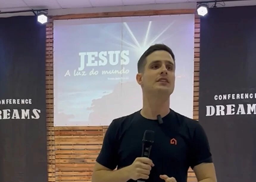 Imagem mostra rapaz discursando na frente de telão onde pode-se ler: Jesus