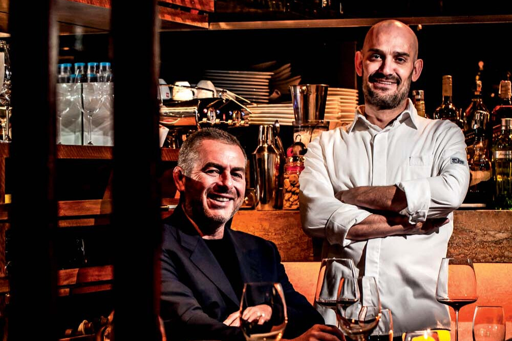 Dois homens com os braços cruzados sorriem para a foto. Um está sentado na frente de uma mesa e usa terno preto. O outro está em pé ao seu lado com avental branco.