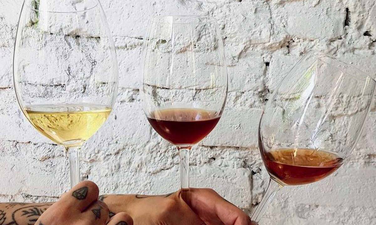 Duas mãos seguram três taças de vinho com cores diferentes. O da esquerda é branco, do meio tinto e da direita alaranjado. O fundo é branco.