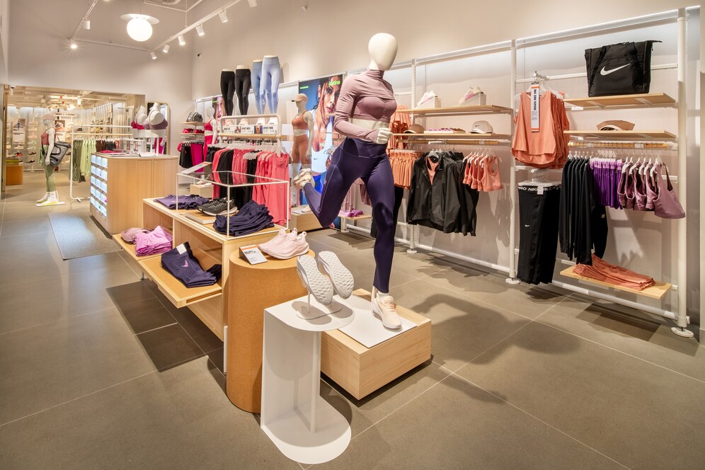 Foto exibe manequim feminino em pose de corrida vestindo calça legging e top. Ao redor, prateleiras com roupas esportivas.
