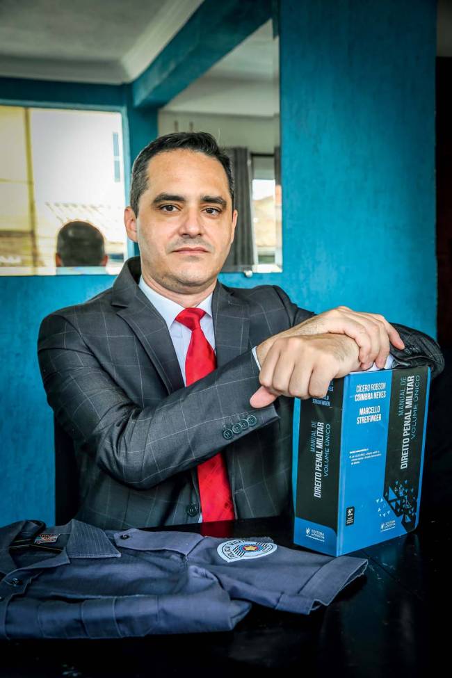 policia militar Imagem mostra Thiago sentado de terno e gravata, com as mãos em cima de um grosso livro de direito militar e, ao lado, uma farda da PM