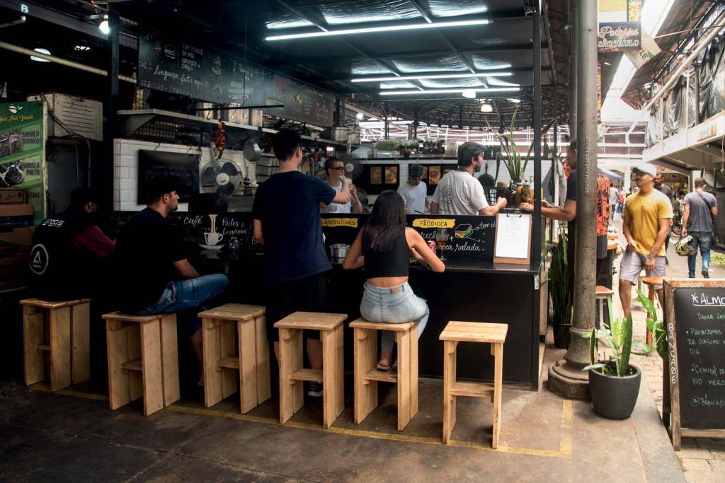 Banca 06 Kinjo Um balcão servindo comida e coberto por cima e aberto dos lados. Uma pessoa está sentada no banquinho de madeira comendo e outras estão na fila fazendo o pedido.