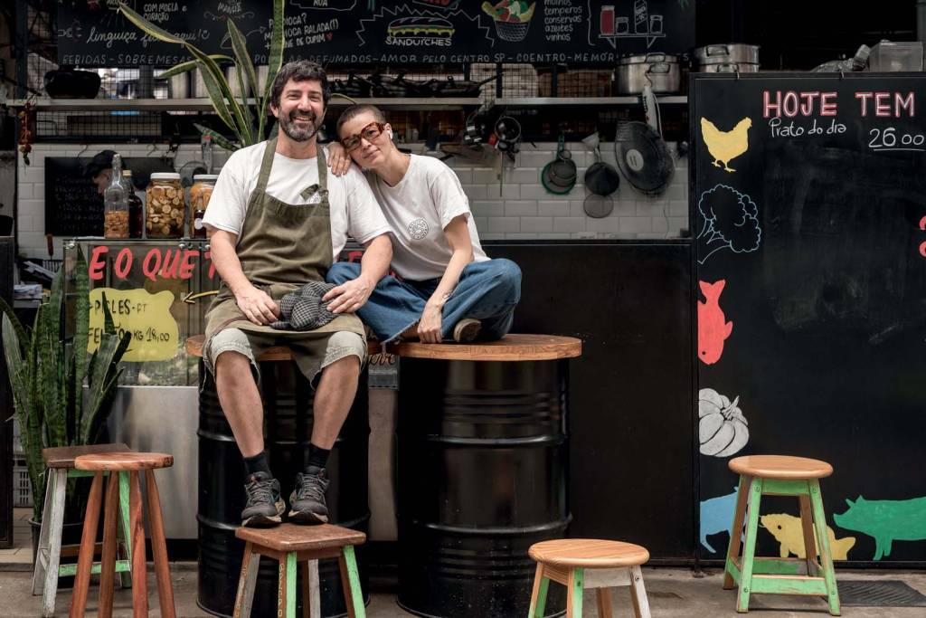 Banca 06 Kinjo O casal posa para a foto, os dois estão sentados em barris e a mulher apoia a mão e a cabeça no ombro do homem.