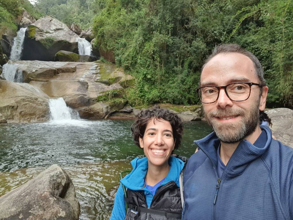 Letícia Alves e Dennis Hyde (Arquivo Pessoal) (4) Casal tira selfie em frente a cachoeira. Ele tem barba e usa óculos de grau, ela cabelos castanhos e posa sorrindo. Ambos vestem paravento azul.
