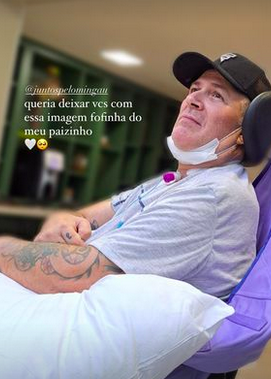 mingau Imagem mostra Mingau sentado, com máscara no queixo. Ele usa boné e está com travesseiro no colo e olha para o canto esquerdo da foto. Ao fundo, uma estante. Texto inserido na foto diz: