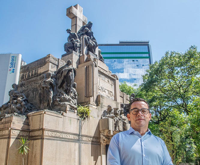 Homem branco de óculos de grau posa em frente a mausoléu a céu aberto, um túmulo alto com cruz no topo e estátuas.