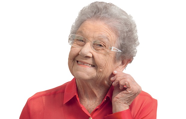 Palmirinha posa com uma mão puxando a orelha, sorrindo e vestindo camisa vermelha de botões dourados.