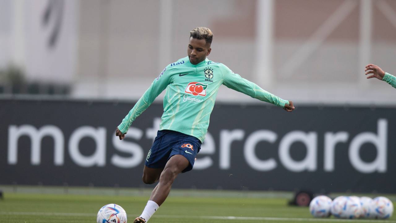 Homem negro chutando bola em campo de futebol. Veste camiseta longa verde com shorts azul
