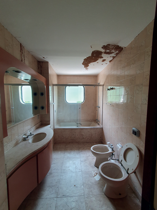 edifício penthouse Imagem mostra banheiro com chão e pia de pedra clara. Ao fundo, boxe com banheira e ao lado, pia com espelho. Acima, teto, com a parte direita, em cima do vaso sanitário e do bidê, com manchas escuras