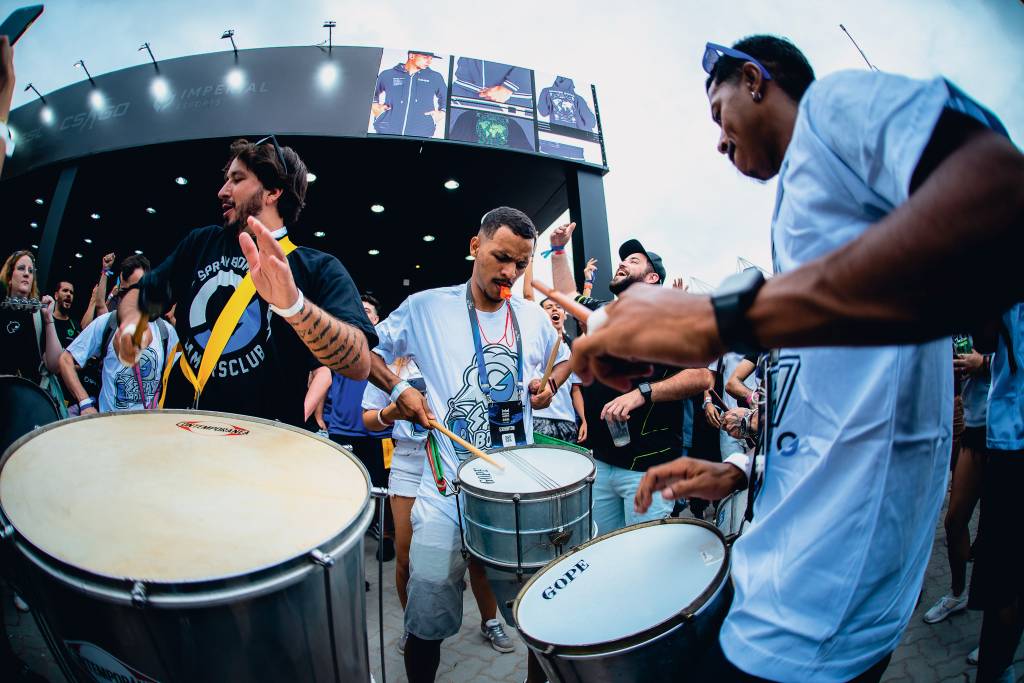 ginásio ibirapuera six invitational rainbow six Imagem mostra bateria tocando em espaço aberto, com três pessoas em primeiro plano, uma com um grande surdo, outra com uma ripa e outra com uma caixa. Todos são homens