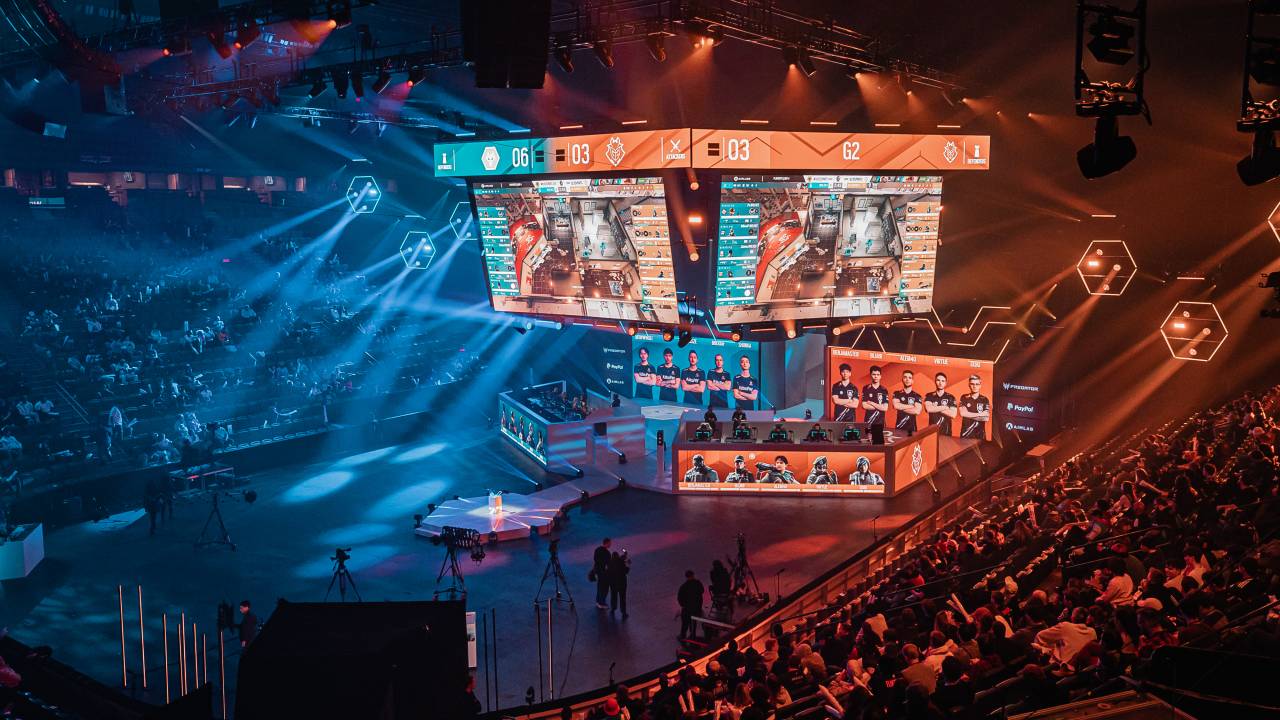 Imagem mostra espaço para eventos escuro, com telões no meio e vários faróis de luzes de iluminação. Embaixo dos telões, duas bancadas, uma azul e outra vermelha, contam com jogadores sentados. Ao redor da imagem, milhares de lugares ocupados por torcedores