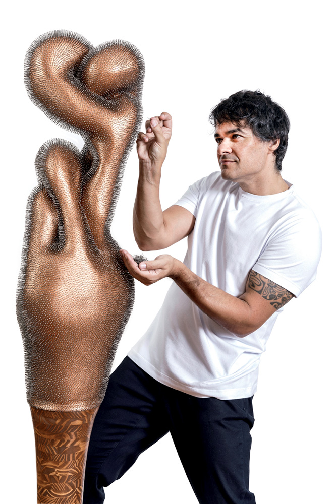 DAN Galeria – Dee Lazzerini – Crédito Edson Kumasaka-Divulgação.jpg Homem branco de camiseta branca e calça preta segura alfinete e o aponta em direção a escultura em tom de bronze