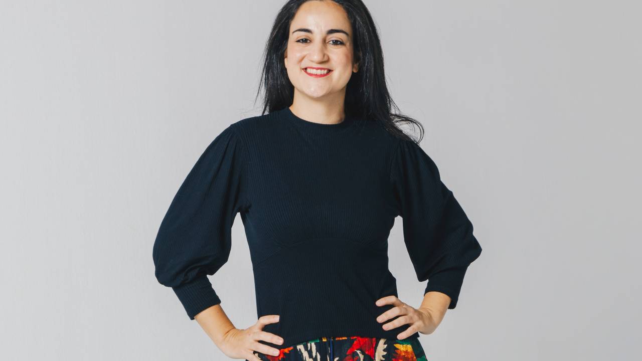 Mulher branca de cabelos longos lisos posa com as mãos na cintura em fundo cinza claro. Veste blusa azul escuro e saia longa estampada