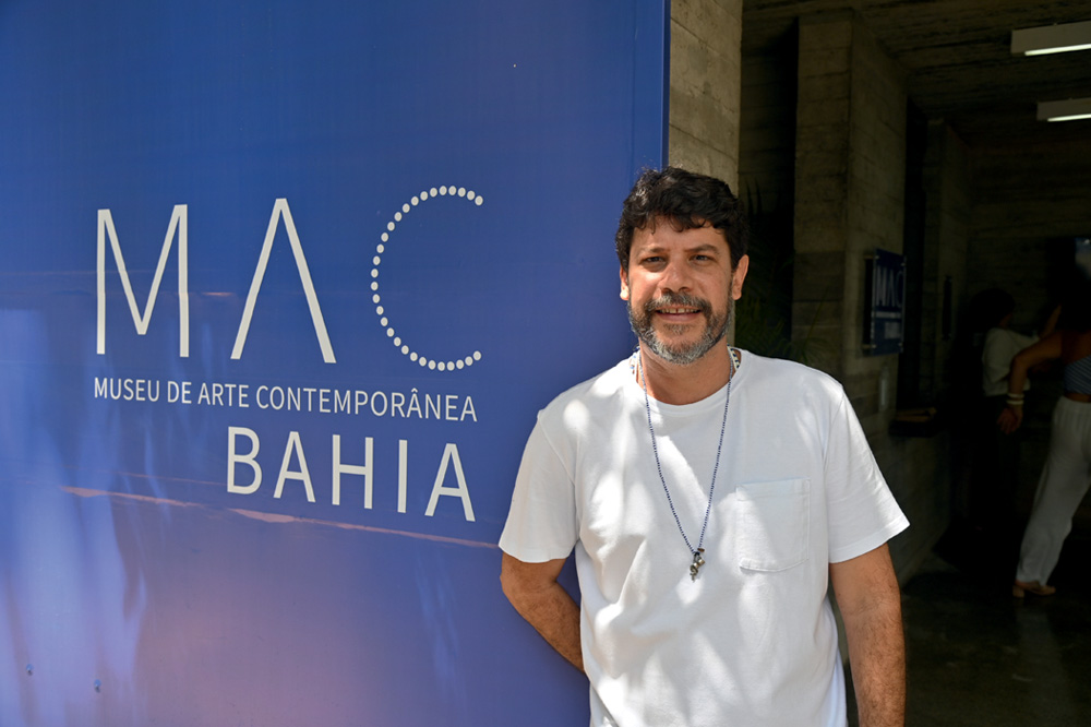 Daniel Rangel (1).JPG Homem branco de camiseta branco ao lado de painel azul indicando MAC Bahia